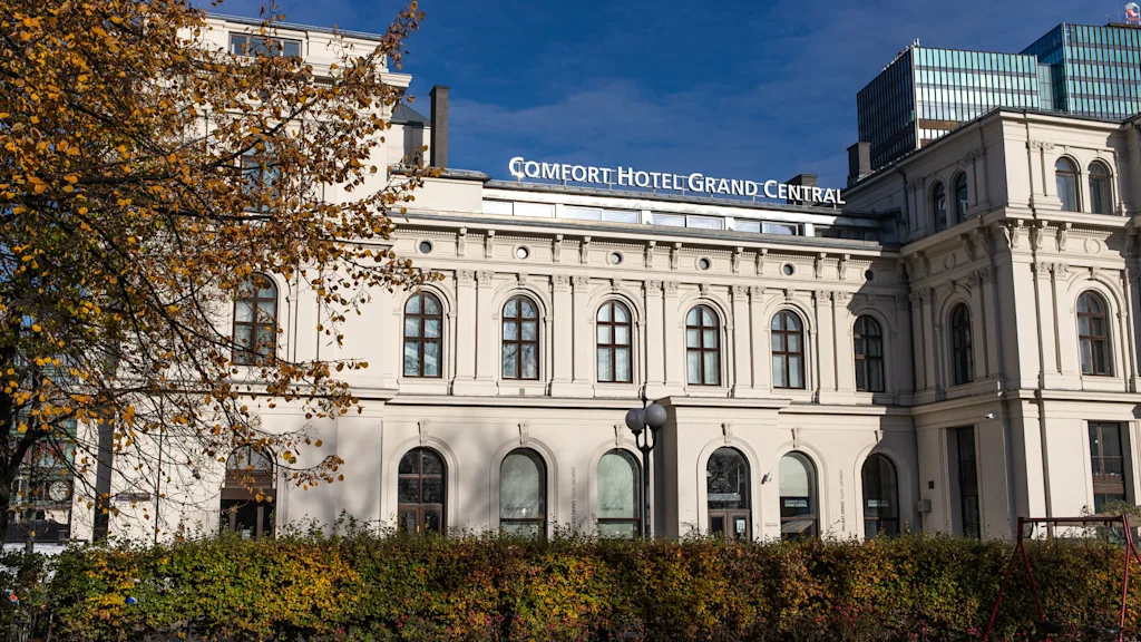 En storslået hotelbygning står omgivet af efterårstræer og buske under en klar blå himmel. Skiltet lyder: "COMFORT HOTEL GRAND CENTRAL."