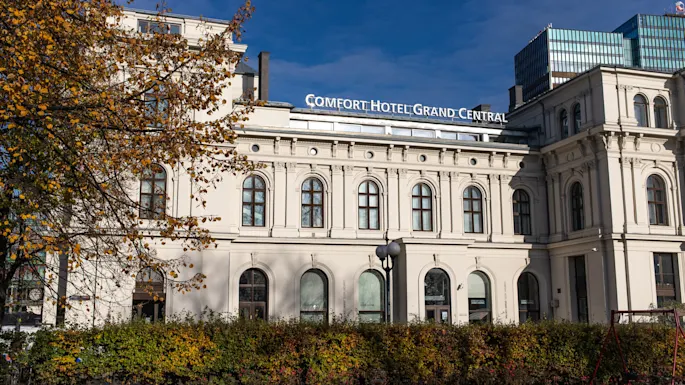 A grand hotel building stands surrounded by autumn trees and bushes, under a clear blue sky. Sign reads: "COMFORT HOTEL GRAND CENTRAL."