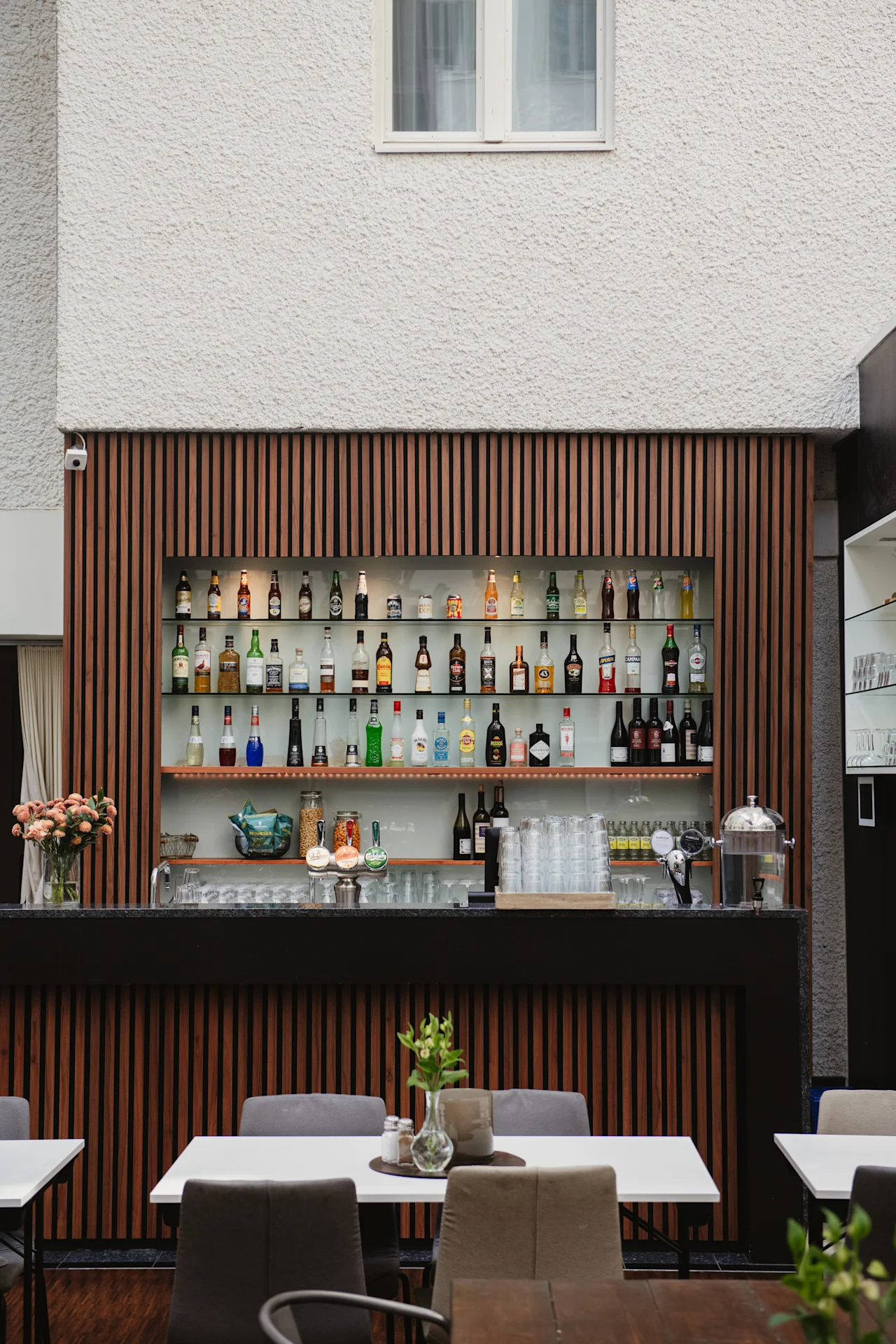 En välfylld bar visar många spritflaskor på hyllor. Placerad mot en vägg med träspjälor, omgiven av bord och stolar, vilket skapar en mysig inomhusmiljö.