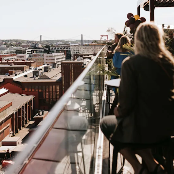 Folk sitter og står på en takterrasse med glassrekkverk, nyter en byutsikt med bygninger og en hengebro. Ballonger og planter gir dekorative elementer.