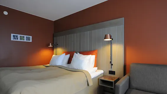 Bed with beige covers and white pillows resting under two wall lamps, next to orange accent cushions. Brown wall with abstract paintings beside a grey sofa.