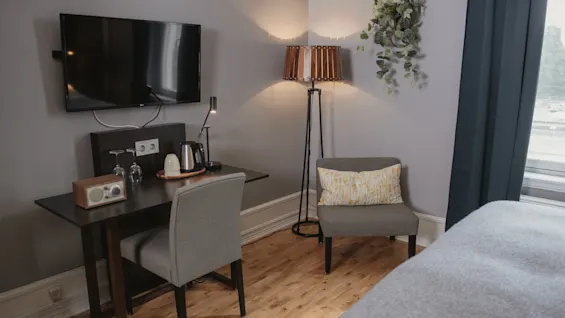 A small desk with a kettle and lamp sits under a wall-mounted TV. Nearby, a chair and standing lamp provide seating. A bed is partially visible.