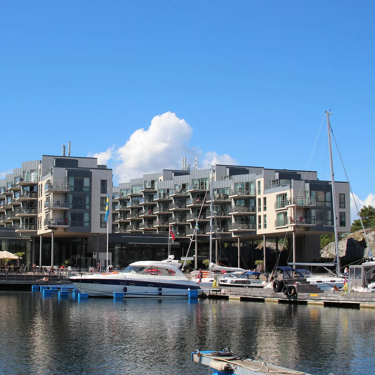 Moderne leilighetsbygninger står ved en marina med flere fortøyde båter. Uteområder med parasoller og flagg ses under en klar blå himmel.