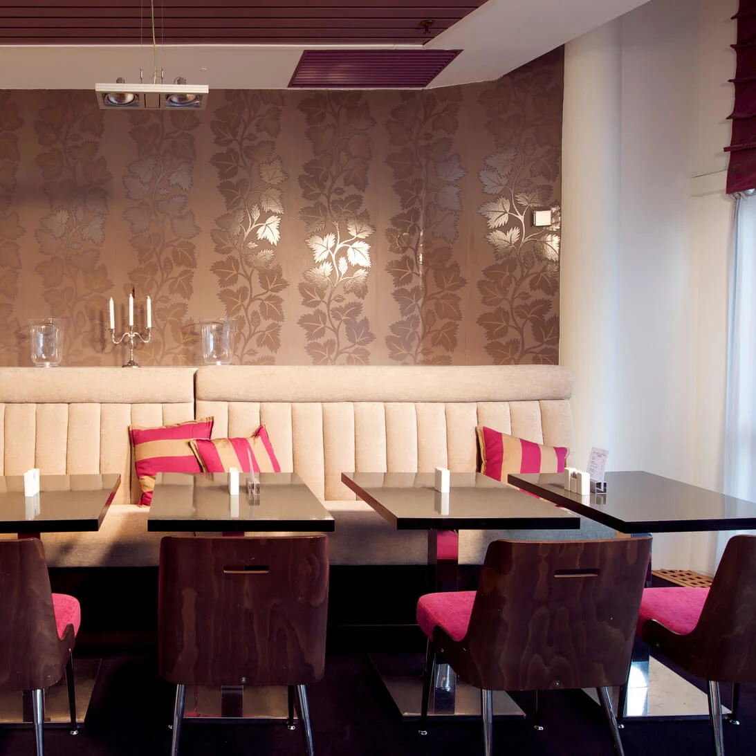 En række moderne, mørke borde og stole vender mod en beige polstret bænk med stribede puder i en stilfuld café, omgivet af mønstrede vægge og elegant dekoration.