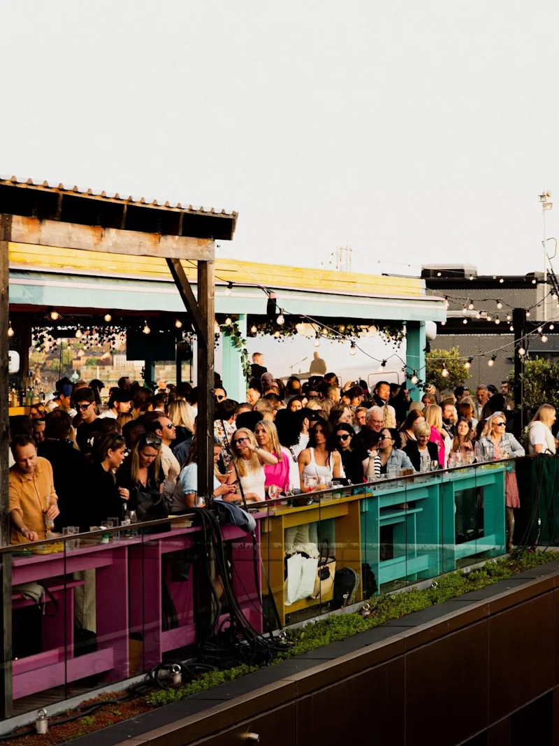 Människor umgås på en färgglad takterrass; många står och pratar vid räcken under ljusslingor, vilket skapar en livlig, festlig stämning.