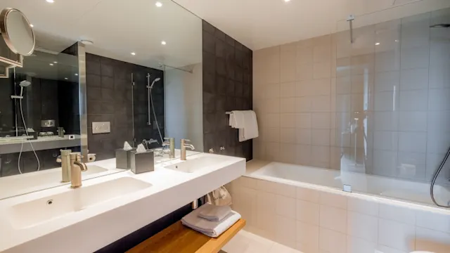 Et moderne bad har en dobbel servant med stort speil, mens et badekar med glasskjerm er ved siden av. Mørke fliser kontrasterer med lysere vegger og gulv.