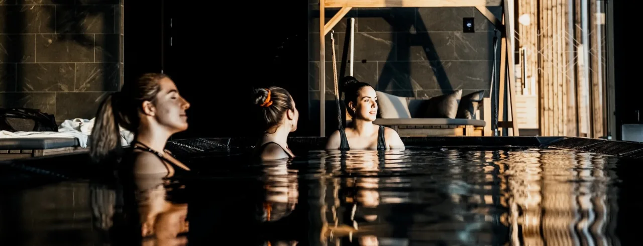 Tre personer befinner sig i en pool, ser avslappnat omkring sig, i en mörk, modern inomhusmiljö med varm belysning.