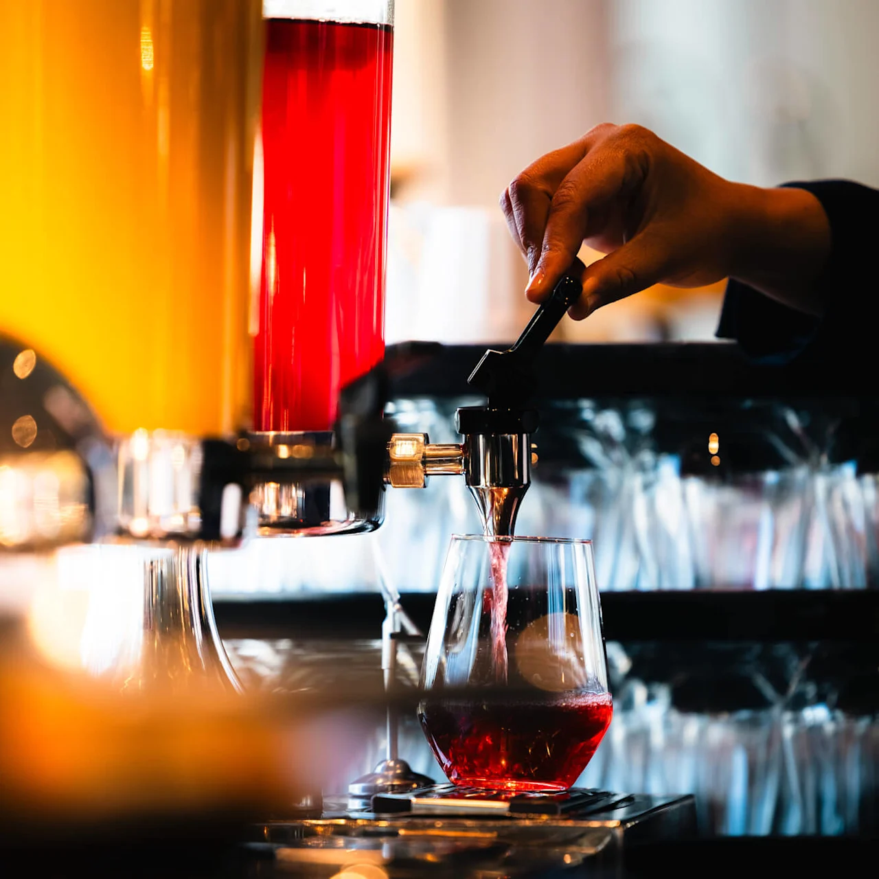 En hand häller upp röd juice från en klar behållare i ett glas, i en ljus miljö med rader av tomma glas i bakgrunden.