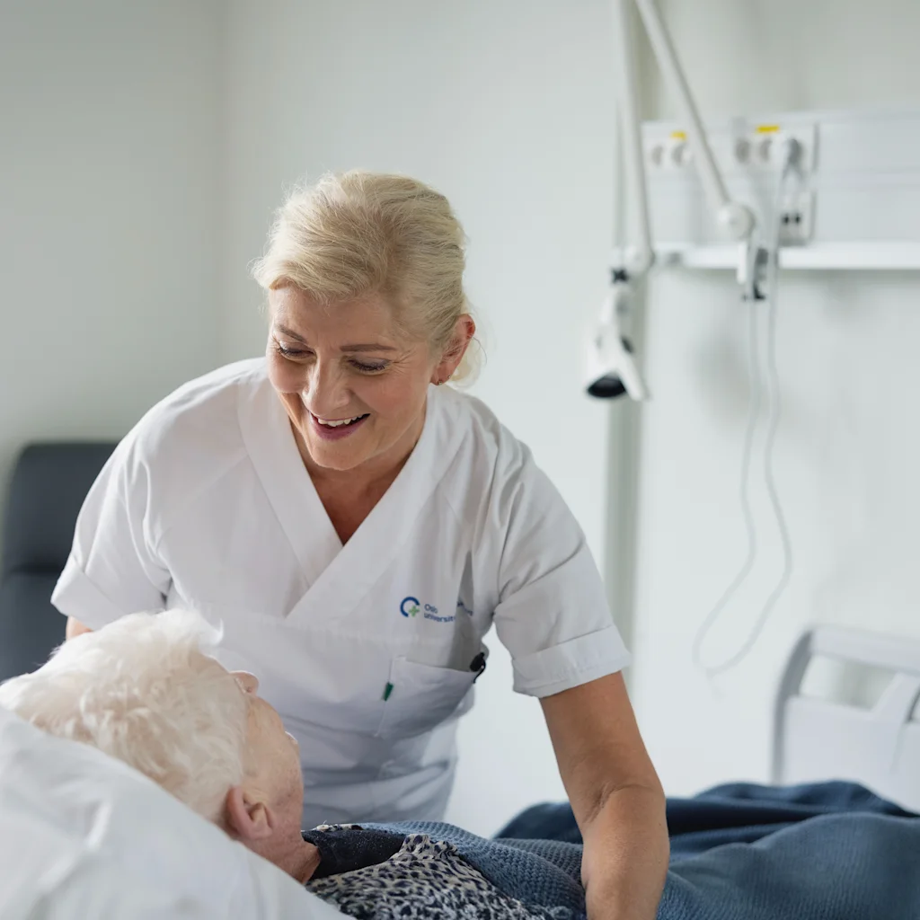 En helsearbeider smiler mens de steller en pasient i en sykehusseng, og viser omsorg og trøst.