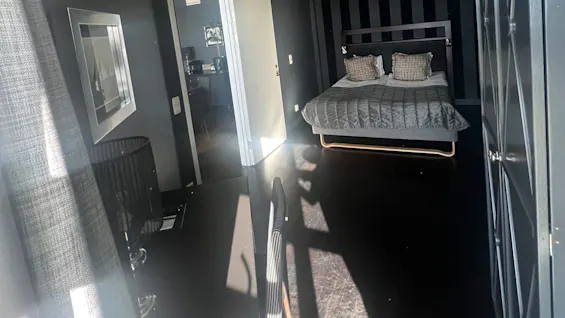 A bed with pillows rests against a striped wall in a dark room. Sunlight streams through a window onto a desk and chair, casting shadows on the floor.