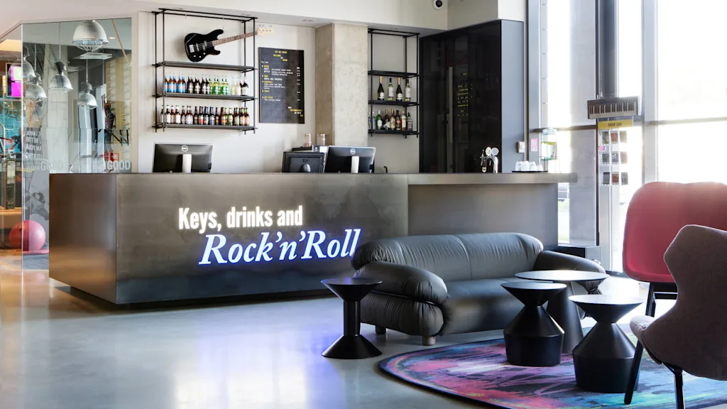 Ett modernt receptionsbord visar texten "Keys, drinks and Rock'n'Roll" med en gitarr och flaskor ovanför. Bekväma sittmöbler och färgglad inredning förstärker den välkomnande loungemiljön.