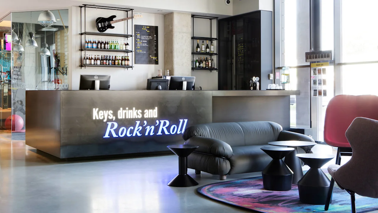 A modern reception desk displays the text "Keys, drinks and Rock'n'Roll" with a guitar and bottles above. Comfortable seating and colorful decor enhance the welcoming lounge atmosphere.