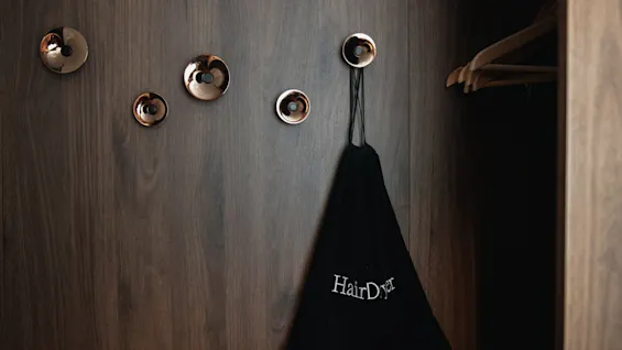 En mørk trevegg med flere runde, metalliske kroker. En svart pose merket "HairDryer" henger fra en krok ved siden av et åpent skap med kleshengere.
