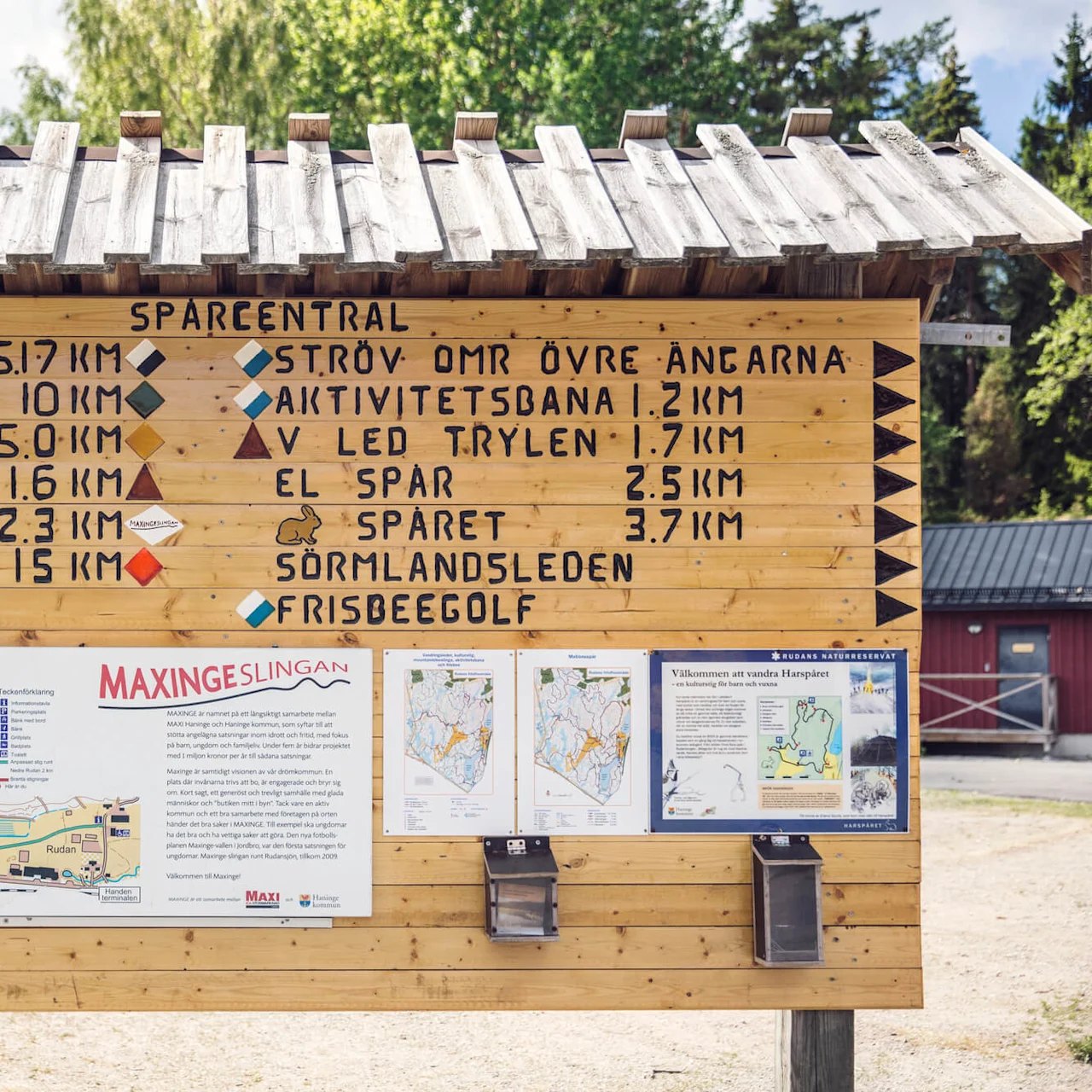 En träskylt visar vandringsavstånd och faciliteter med kartor och broschyrer i en skogsmiljö. Texten innehåller: “SPARCENTRAL”, olika spårnamn, avstånd och ikoner för aktiviteter.