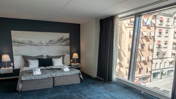 Hotellrum med en prydligt bäddad säng med grå och vita sängkläder, flankerad av sängbord med lampor. Stort fönster med utsikt över en stadsgata med klassiska byggnader.