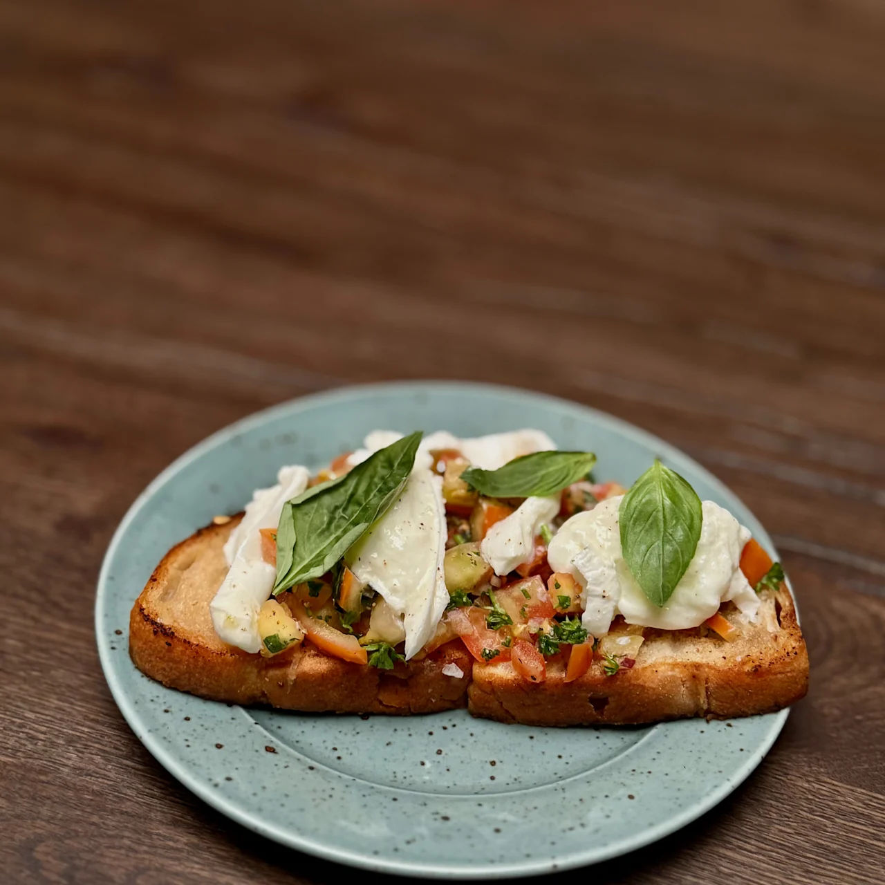 A slice of toasted bread topped with diced tomatoes, mozzarella slices, and basil leaves rests on a speckled green plate, placed on a wooden table.