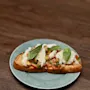 A slice of toasted bread topped with diced tomatoes, mozzarella slices, and basil leaves rests on a speckled green plate, placed on a wooden table.