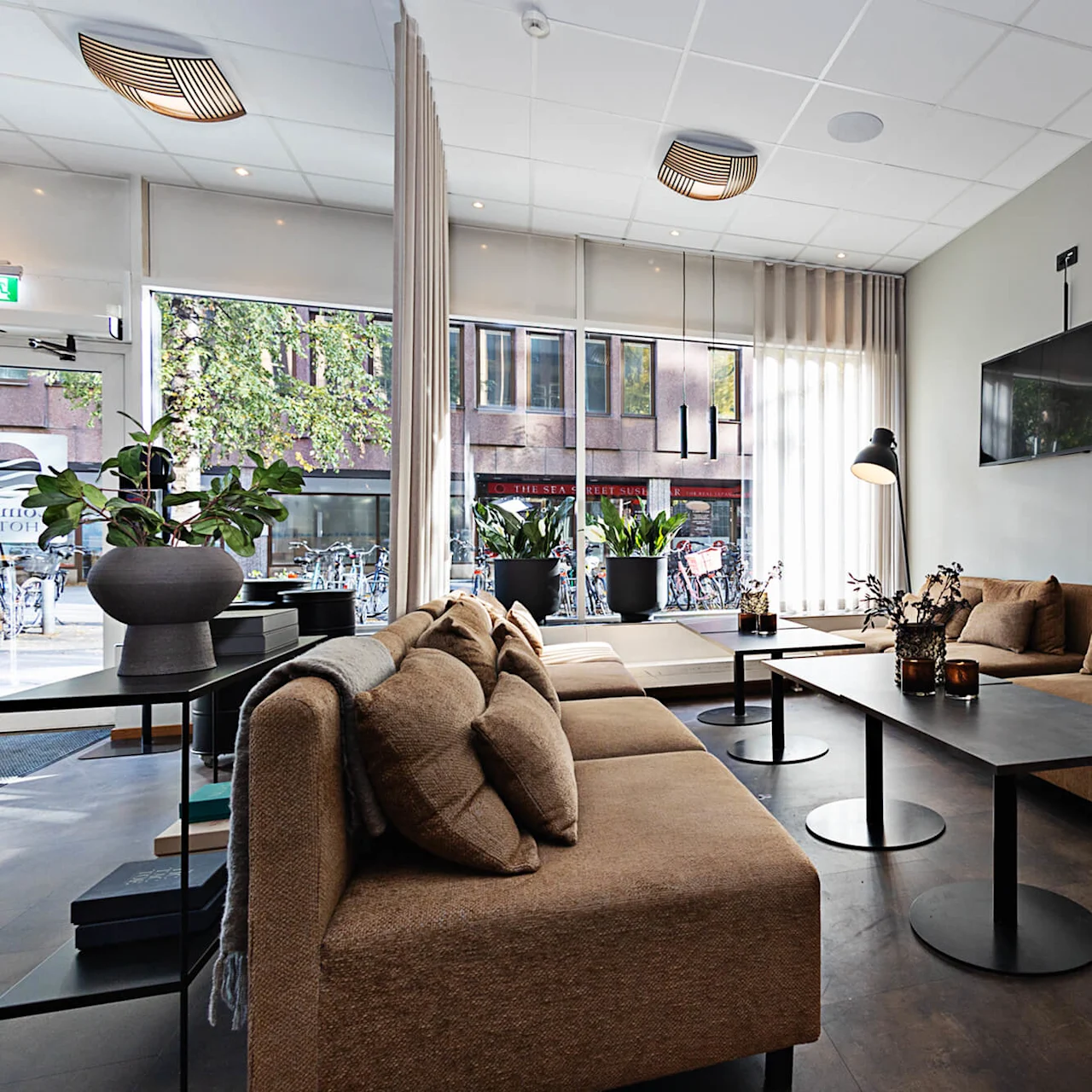 Bruna soffor formar loungens moderna stil och vätter mot en elegant svart TV på väggen. Höga fönster visar en stadsgata utanför, vilket förstärker naturligt ljus och atmosfär.