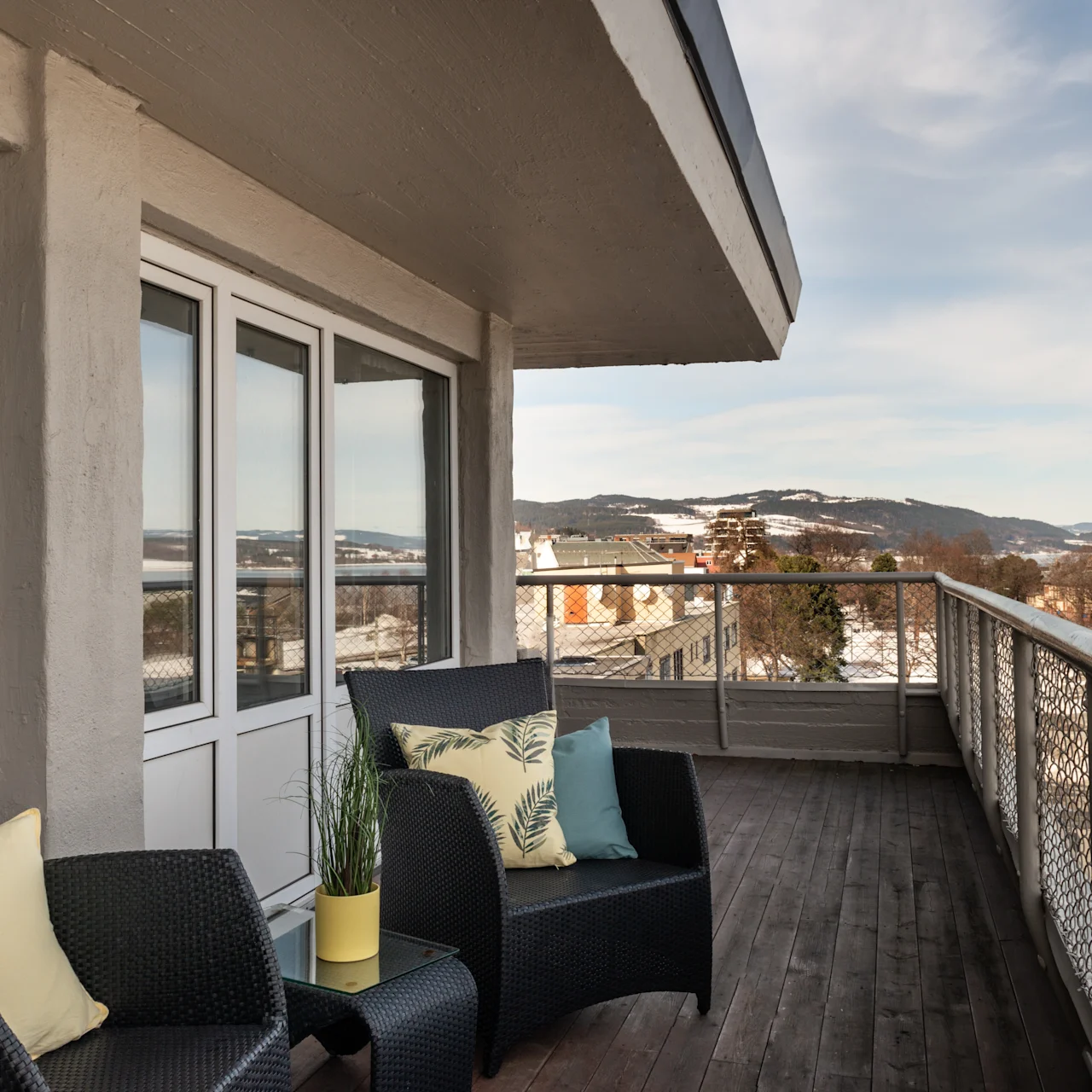 Outdoor balcony set with wicker chairs holding pillows, situated by a glass table with a plant. Overlooks snow-covered rooftops and distant mountains under a cloudy blue sky.