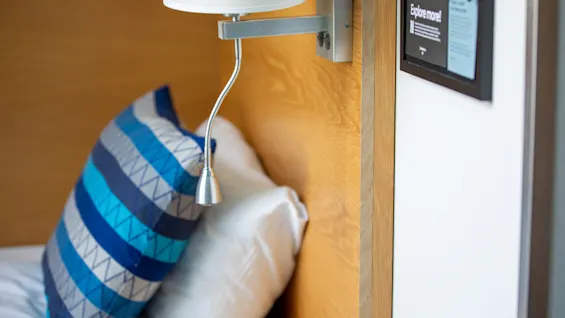 En vegglampe med fleksibel hals lyser opp en seng med blåstripete puter mot en trehodegavel. I nærheten står et skilt med teksten "Explore more!".