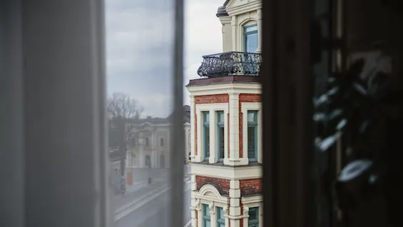 A red brick and cream facade building is viewed through a window, with black ornate balcony railings and an overcast sky in the background.