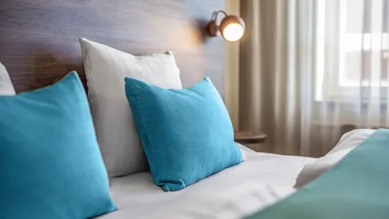 A cozy bed features vibrant blue pillows and a neatly arranged white duvet, under soft, warm lighting beside a wooden headboard in a well-lit room with sheer curtains.