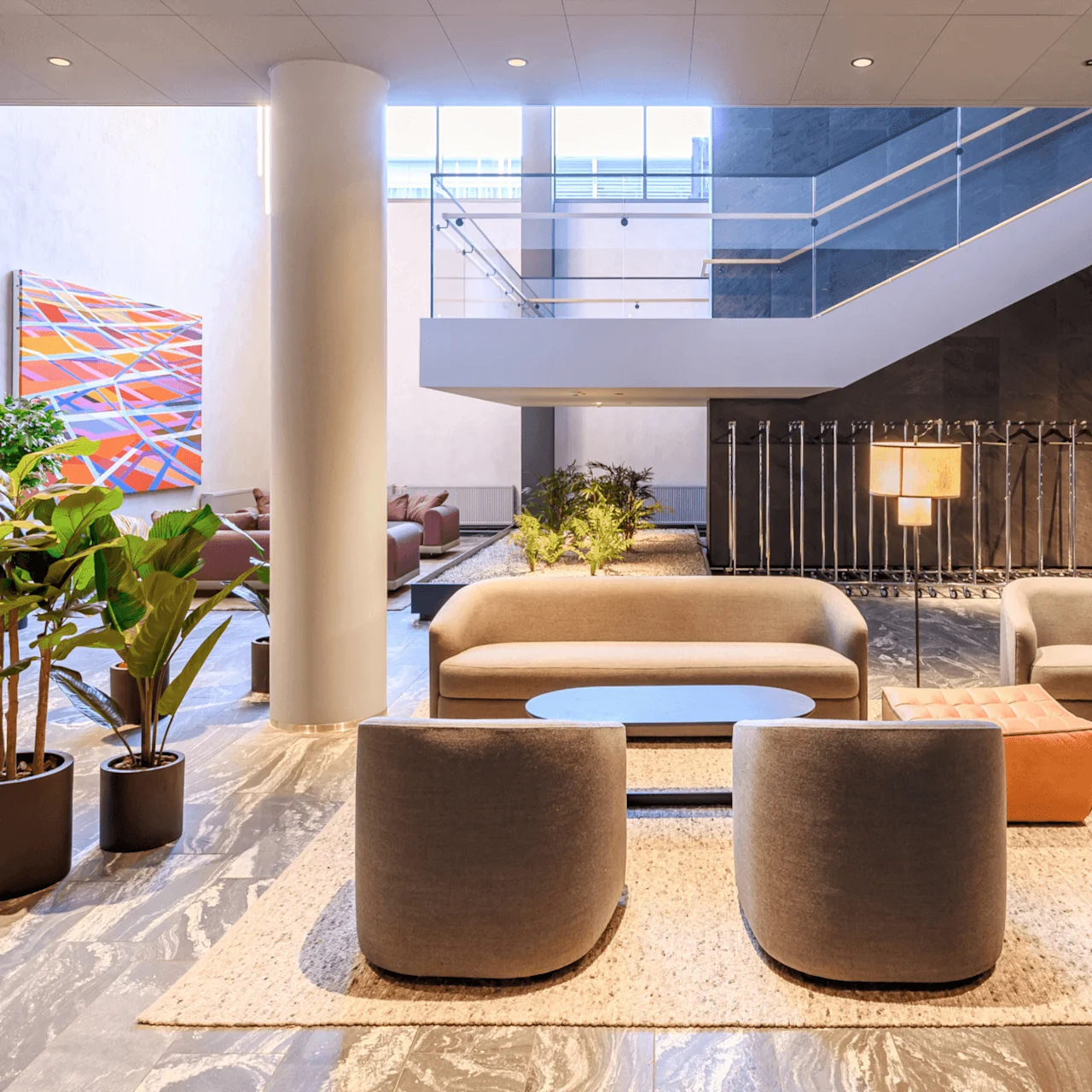 A modern lobby features gray armchairs and a sofa around a table on a rug, with potted plants nearby. A large staircase and abstract art piece complement the spacious design.