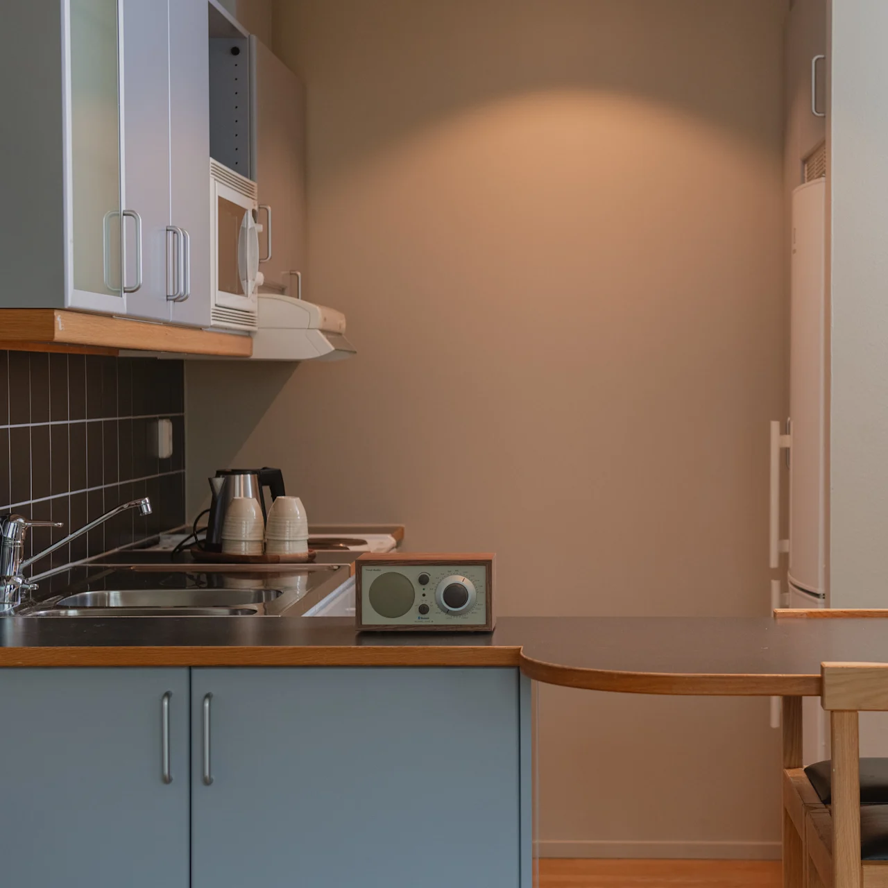 Ett litet kök med ljusblå skåp, en diskho och en bänkskiva med en radio och två vattenkokare. Ett matbord med stolar är delvis synligt. Beige väggar omger.