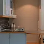 Ett litet kök med ljusblå skåp, en diskho och en bänkskiva med en radio och två vattenkokare. Ett matbord med stolar är delvis synligt. Beige väggar omger.