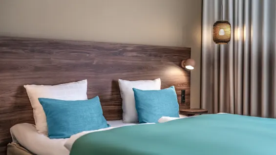 A bed with teal pillows and blankets sits against a wooden headboard. A small wall lamp and hanging light illuminate the bedroom, with beige curtains on the right.