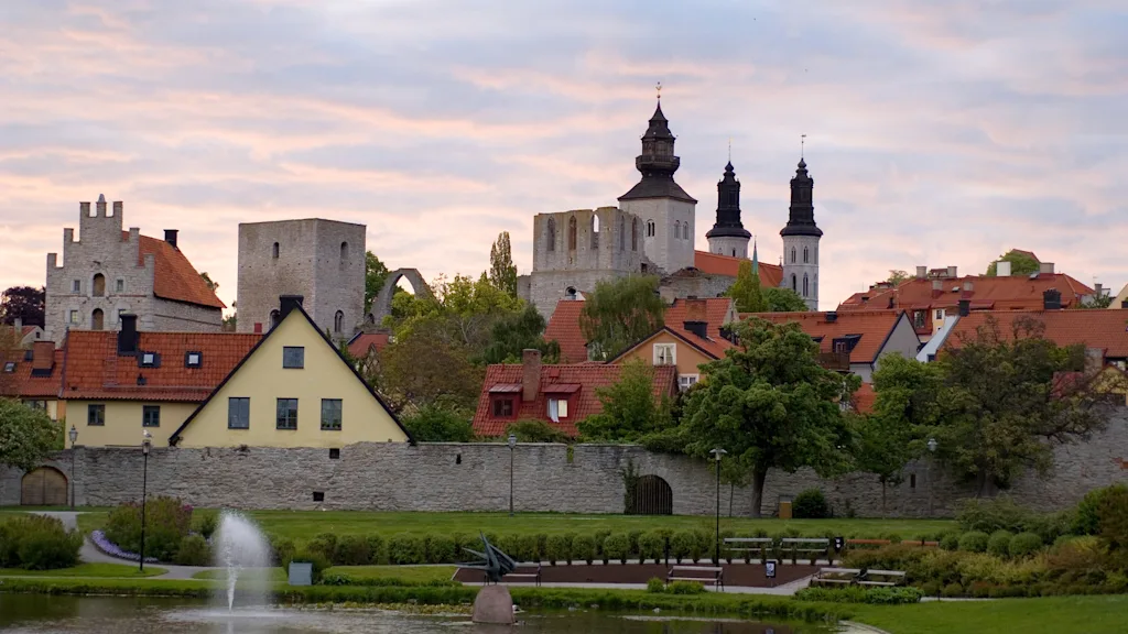 En medeltida kyrka reser sig över en stad med färgglada byggnader. En stenmur och lövträd omger området. En fontän sprutar vatten i en närliggande parkdamm.