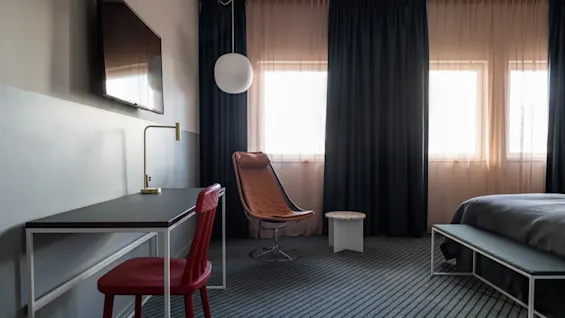 Television mounted on the wall, above a desk with a red chair, in a hotel room with a bed, large windows, blue curtains, and modern furniture. 
