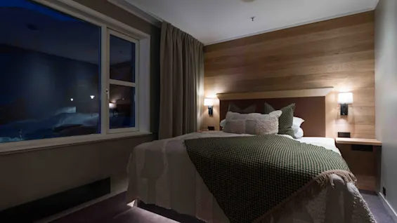 A neatly made bed with patterned pillows and a blanket, positioned against a wooden panel wall with lamps, is accentuated by window curtains; visible outside is a dark, snowy scene.