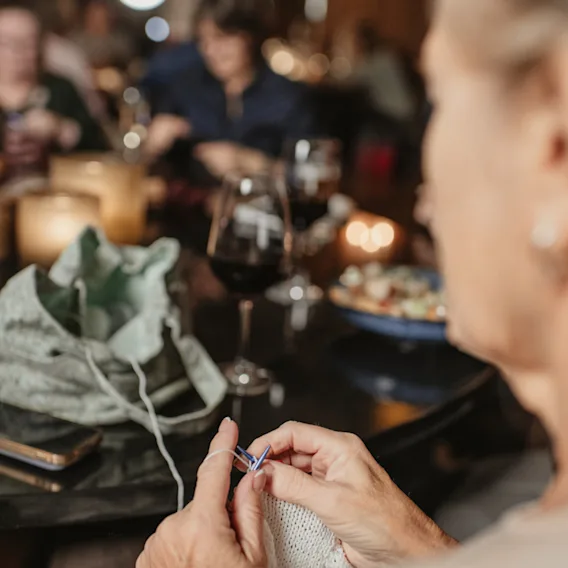 En person som strikker med garn og pinner i et dempet sosialt miljø, med drinker og andre mennesker i bakgrunnen.