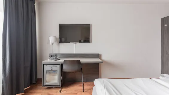 A hotel room featuring a desk with a TV mounted above it, a mini-fridge, a chair, and a bed with white linens. Dark curtains are drawn on the left.