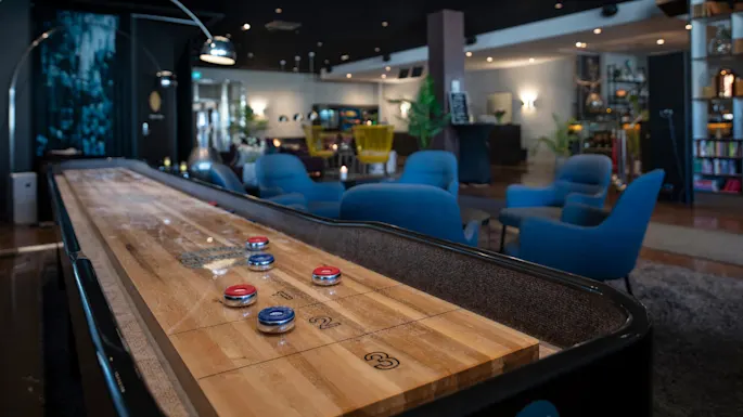 Et shuffleboard-bord med røde og blå brikker står i en koselig lounge med blå stoler, planter og hyller. Tekst: "BEWARE Mind your fingers."
