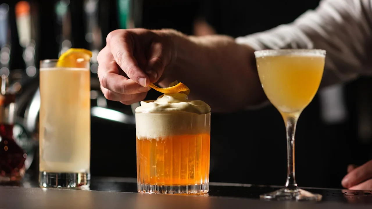 En hand dekorerar en orange cocktail med skum i en svagt upplyst bar, omgiven av andra cocktails.