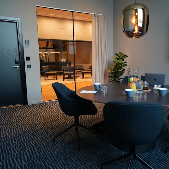 A round table with four chairs holds cups and a laptop, under a hanging light, in a meeting room with a glass wall revealing a lounge outside.