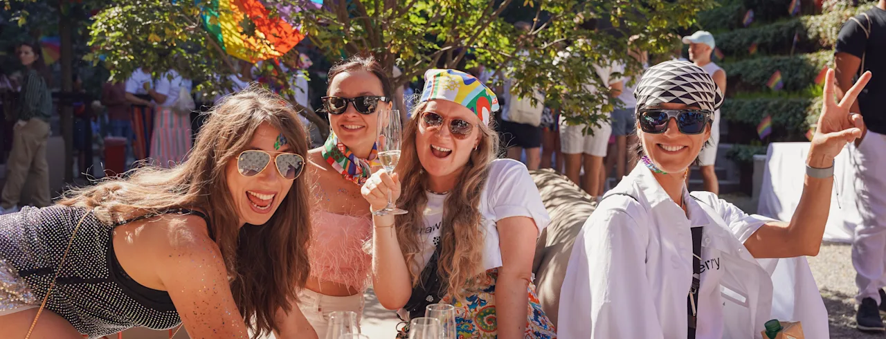 Four joyful people celebrating outdoors with drinks and rainbow decorations, one making a peace sign.