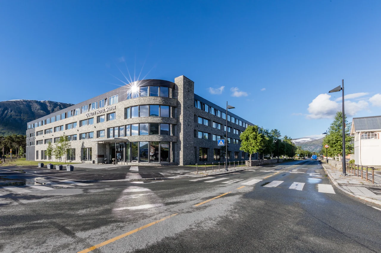 En moderne fleretagers bygning med navnet "Scandic Valdres Hotel" står på et gadehjørne, med reflekterende vinduer omgivet af træer, bjerge og en klar blå himmel.