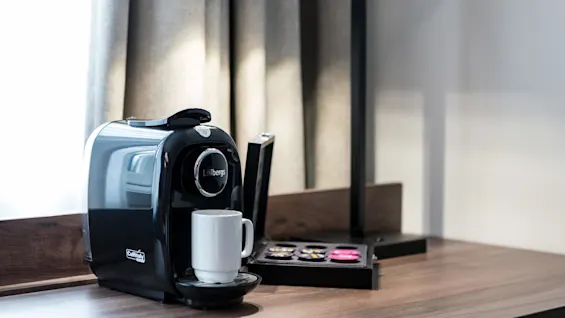 A black Löfbergs coffee machine rests on a wooden countertop, brewing into a white mug. Nearby, there are assorted coffee pods under a modern lamp, in a softly lit room.