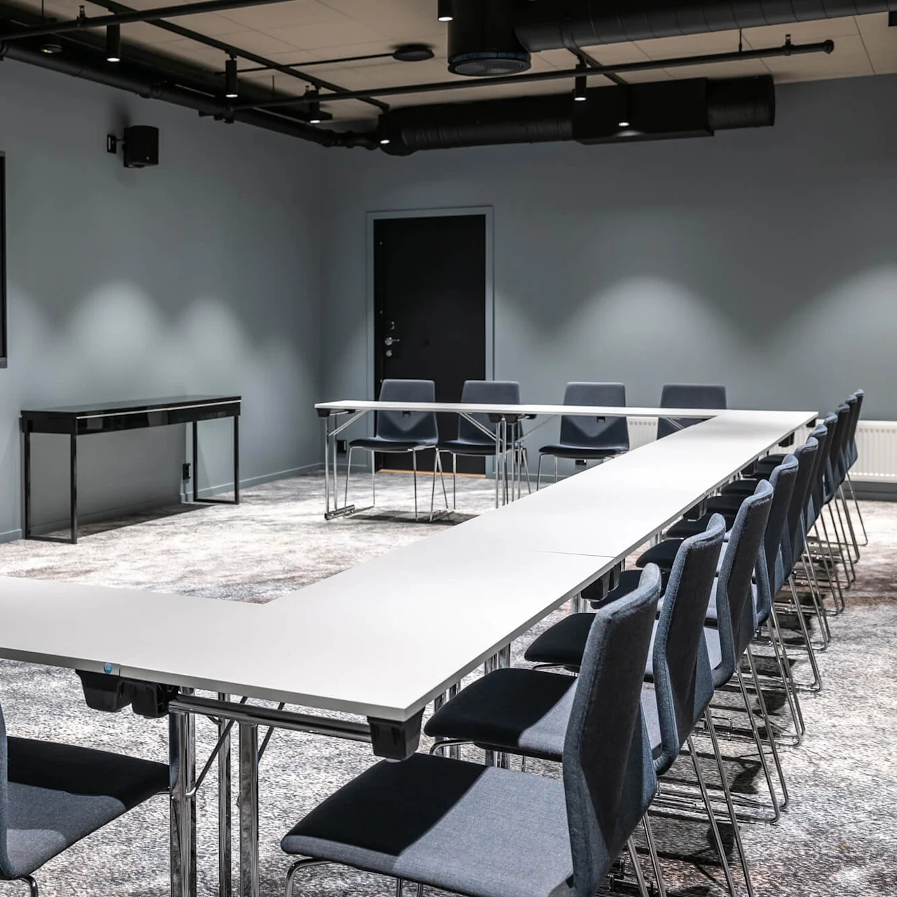Et stort U-formet konferencetable er omgivet af flere stole i et tæppebelagt rum, med en vægmonteret skærm og minimalistisk dekoration.