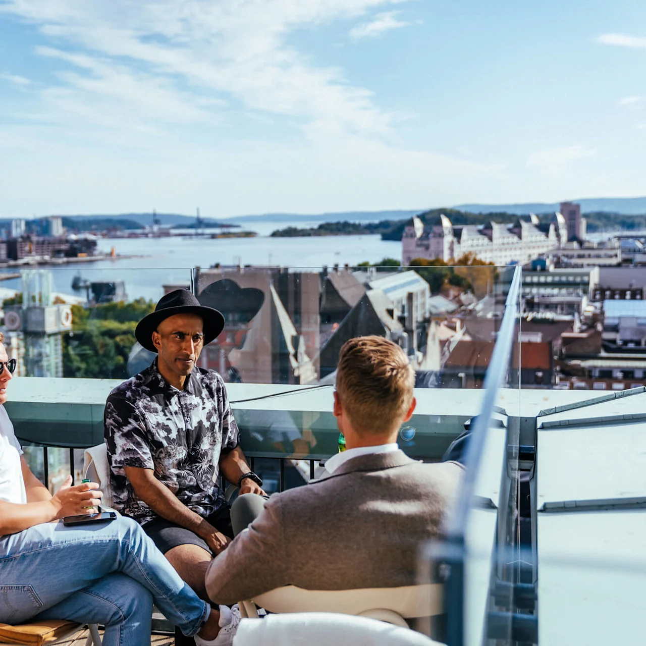 Three people sit conversing on a rooftop terrace overlooking a city with a river in the background on a sunny day.