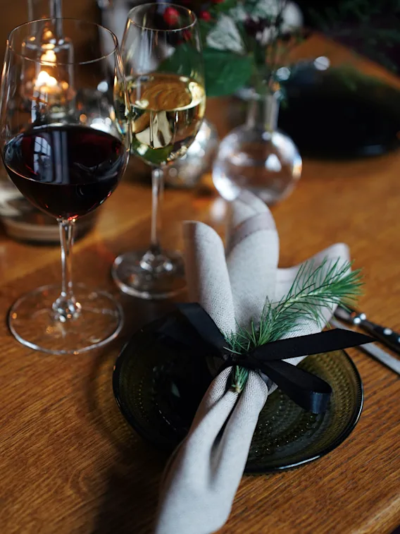 Festive dinner table setup with red and white wine glasses and a cloth napkin tied with a black ribbon and pine sprig on a green plate.