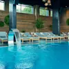 A luxurious indoor pool features water cascading from steel fountains. Surrounding the pool are wooden lounge chairs with cushions, lush greenery, and large, decorative wall accents.