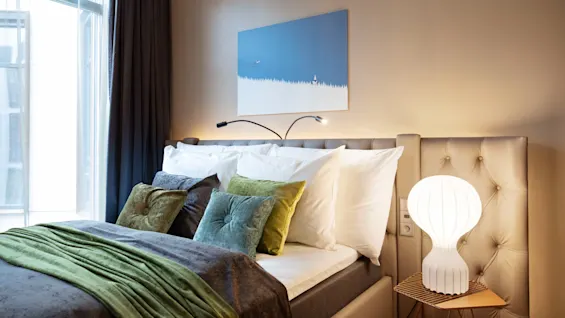 A bed with green and gray bedding rests against a tufted headboard. A modern lamp sits on a small table, beneath a blue and white painting. Sunlight filters through a window.