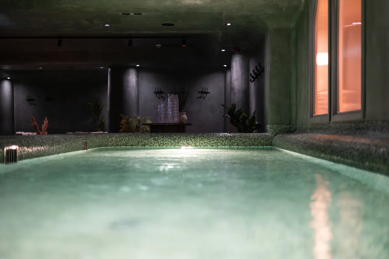Hämärästi valaistu sisäuima-allasalue vihreillä laatoitetuilla reunoilla, kasveilla ja ikkunalla, mikä viittaa rauhalliseen kylpyläkokemukseen.