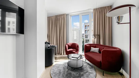 Red sofa and chair sit near a round, glass coffee table on a gray rug. Sunlight streams through floor-to-ceiling curtains, brightening this modern room with a wall-mounted television.
