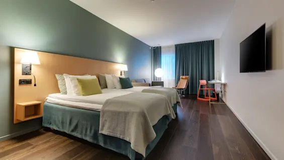 En pent redt seng med puter og tepper, ved siden av en tent vegglampe, i et moderne hotellrom med tregulv, et skrivebord med en oransje stol og et stort vindu med gardiner.