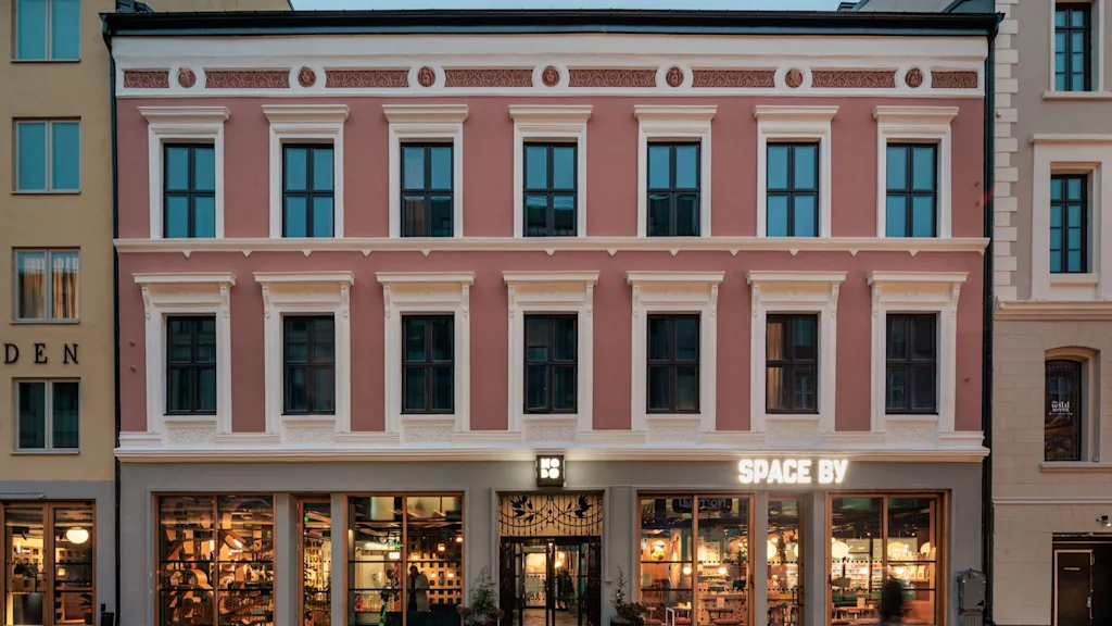 A pink building with white trim and many windows. The ground floor has large illuminated display windows and an entrance. Text reads SPACE BY.